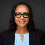 Professional headshot of a smiling Black woman with braided hair, wearing black-framed glasses, a light blue blouse, and a black blazer against a dark backdrop.