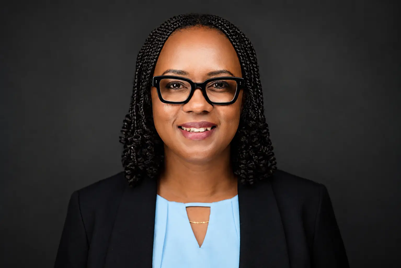 Professional headshot of a smiling Black woman with braided hair, wearing black-framed glasses, a light blue blouse, and a black blazer against a dark backdrop.