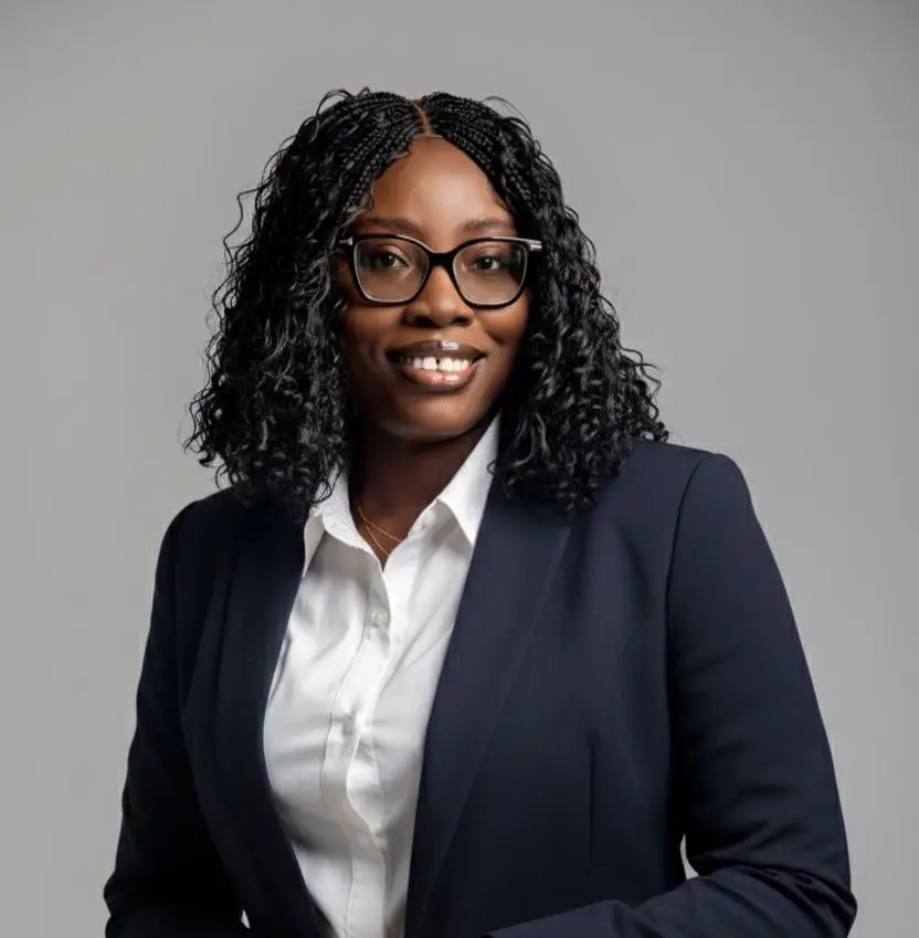 Professional portrait of a smiling Black woman wearing glasses, a white shirt, and a navy blazer against a gray background