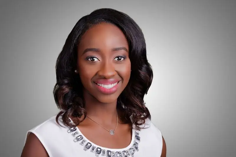 Portrait of a smiling woman with shoulder-length wavy hair wearing a white top with a jeweled neckline against a neutral gray background, in a professional headshot style