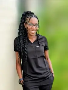 Smiling woman in black scrubs with long braids and glasses leaning against a light wall outdoors.